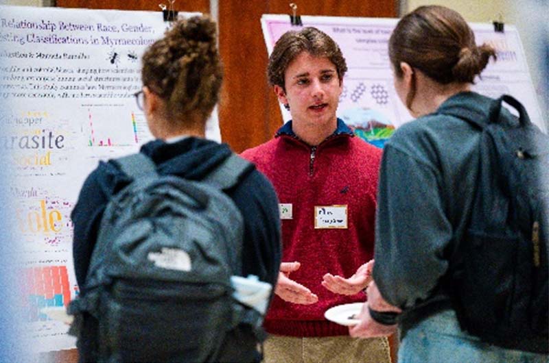 A man with a nametag is standing in front of his poster board explaining it to two women