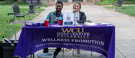 About thumbnail - 2 people sitting behind a table that says WCU