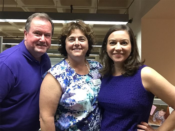 Color photograph of three adults standing close together indoors and smiling at the camera. The person on the left wears a purple polo shirt, the person in the center wears a patterned sleeveless top with a name badge, and the person on the right wears a sleeveless purple shirt.