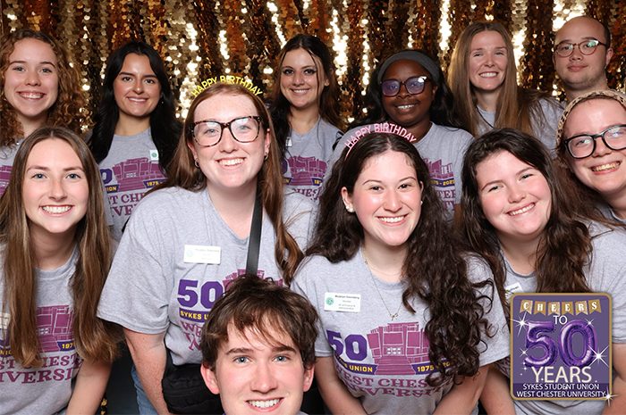 A group of student staff members pose together and smile at the camera in front of a gold sequin backdrop. They wear matching gray T-shirts celebrating the 50th anniversary of the Sykes Student Union at West Chester University, and several wear “Happy Birthday” headbands. A small graphic in the corner reads “Cheers to 50 Years — Sykes Student Union, West Chester University.”