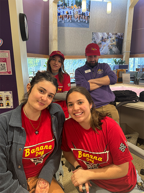 Four student staff members gathered at the information desk in Sykes Student Union. Three individuals wear matching red “Banana Day” T-shirts and smile toward the camera, while two wear red baseball caps with a banana logo on the front. A fourth person stands behind the desk wearing a purple hoodie and cap with arms crossed. Posters, signage, and computer equipment are visible around the desk.