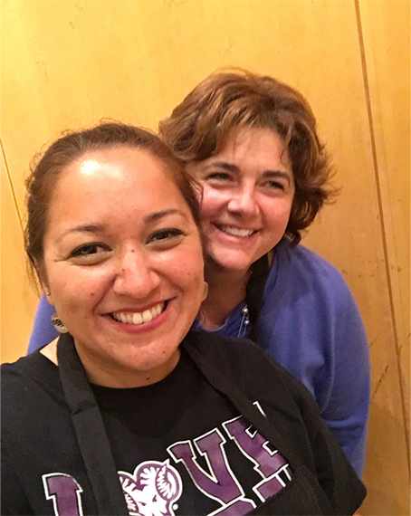 Color photograph of two people smiling at the camera indoors. One person stands slightly in front wearing a dark T-shirt with university lettering, while the other stands just behind wearing a blue top.