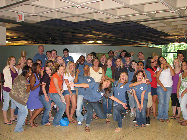 A large group of students and staff pose together indoors during a staff training event. The group stands closely packed, smiling, laughing, and making playful gestures toward the camera. Several people wear matching blue T-shirts with name tags, while others wear casual summer clothing.