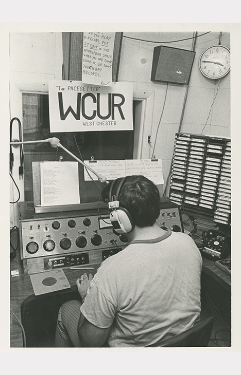 Black-and-white photograph of a student seated at a radio broadcast console inside a campus radio station studio. The student wears large over-ear headphones and faces audio equipment with knobs, switches, and microphones. A sign above the window reads “The Pacesetter WCUR West Chester.” A wall clock, speakers, handwritten notes, and a rack of cassette tapes are visible