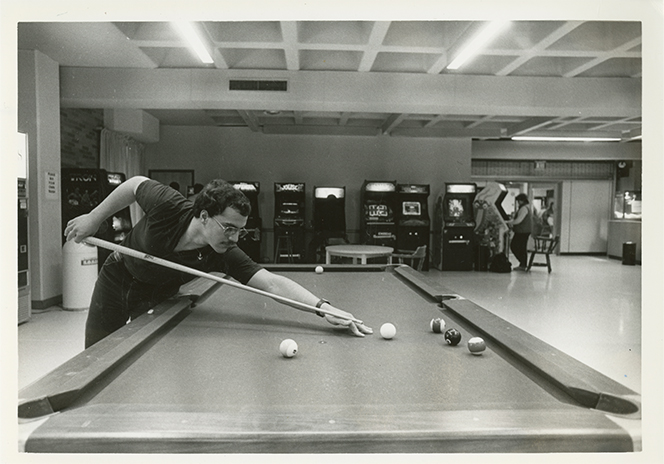 A black-and-white photograph of a student leaning over a pool table in a college union game room, aiming a shot with a cue stick. Several billiard balls are spread across the table. In the background, rows of arcade machines line the wall, and other students stand and play games. The space has a low ceiling with recessed lighting.