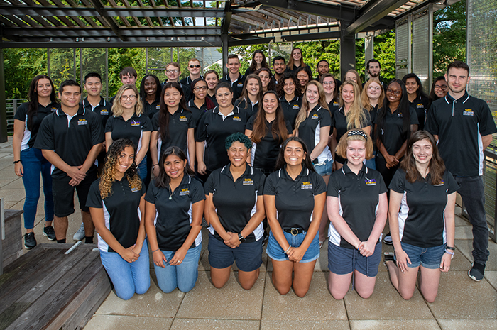 A large group of student staff members pose together outdoors under a covered structure. They wear matching black polo shirts with embroidered logos and stand in several rows, smiling toward the camera. The picture is set on the pergola of Sykes Student Union.