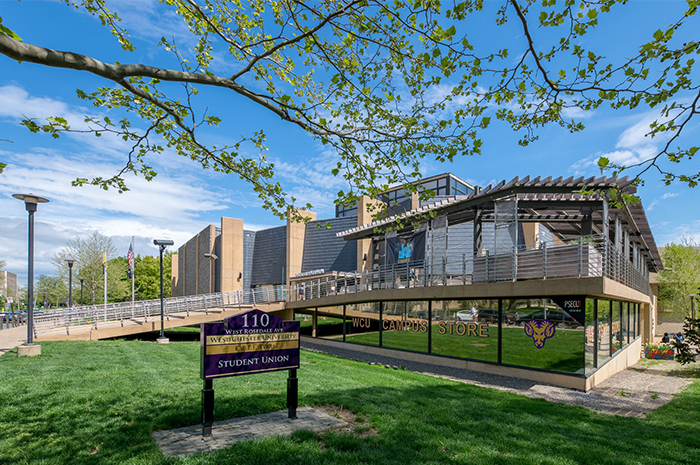 Exterior of the Sykes Student Union on a sunny day. The building features modern architecture with wood accents, metal railings, large windows, and an elevated walkway leading to the entrance. A sign in the foreground reads “110 W. Rosedale Ave — West Chester University Student Union,” and storefront windows display “WCU Campus Store” signage. Trees with spring foliage and a grassy lawn surround the building under a blue sky.