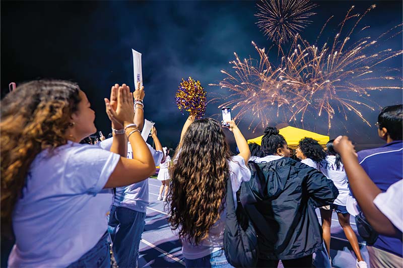 A group of students gathered at Ram Rally Under the Lights taking photos of the fireworks going off in the background