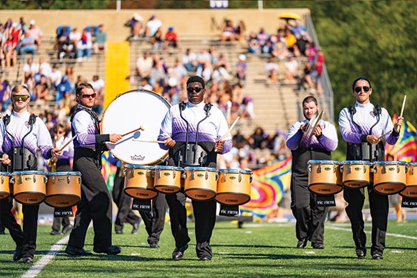 The marching band drum section plays their instruments as the out of focus crowd in the bleachers watches from the back