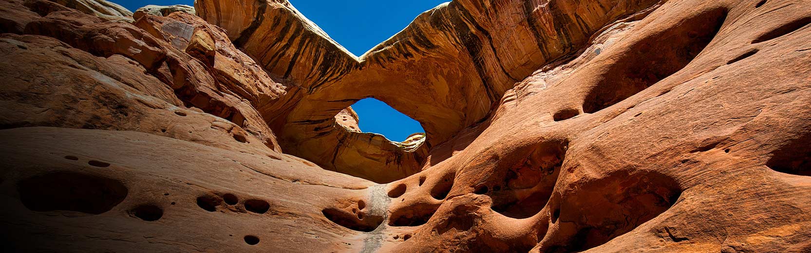 Upward view of a red sandstone rock formation with a natural arch and weathered holes, set against a clear blue sky.