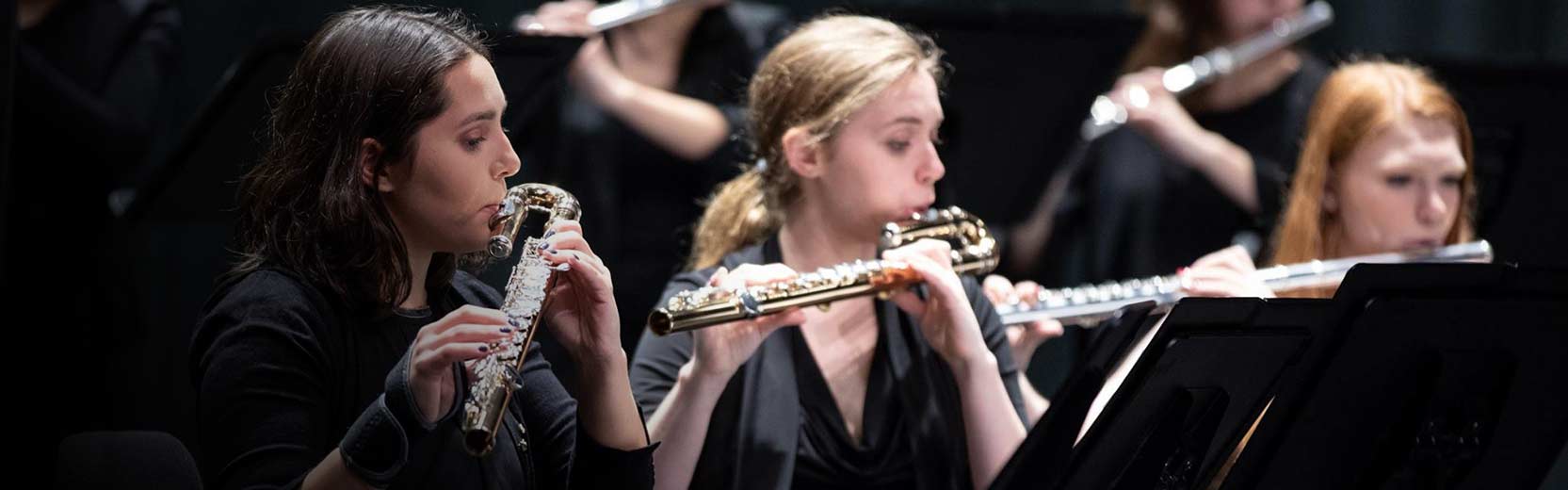 Musicians in a concert ensemble play flutes while reading from sheet music during a performance.